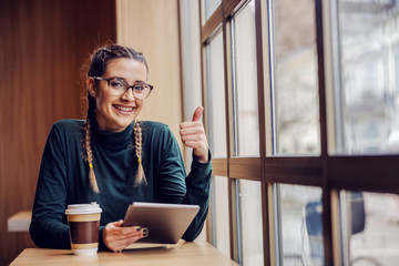 Young smiling geeky girl sitting in coffee shop, enjoying free time, holding tablet and showing thumbs up. New technologies concept.