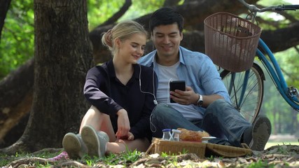 Young couple in love sitting on mat using smartphone listening to the music sharing by headphone on picnic in the park. dating, romance outdoors