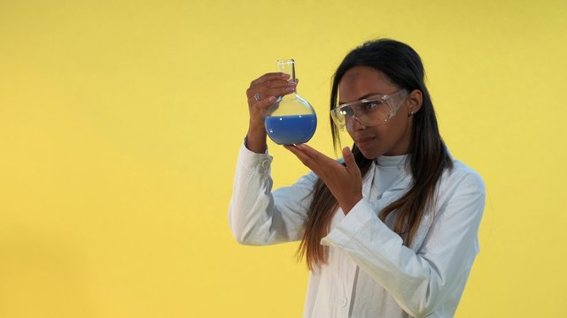 Pretty Black Woman In Safety Glasses And Lab Coat Holding Flask With Liquid In Her Hands. There Is Yellow Background.