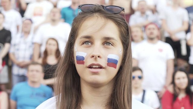 Portrait of Pretty Female football fan sings Russian national anthem cheering at match, supporting football team. Slow motion