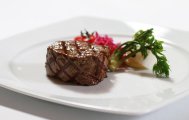 Closeup image of thick juicy steak with grill marks on a white plate with vegetables garnish.