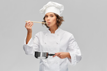 cooking, culinary and people concept - female chef in toque with saucepan and spoon tasting food over grey background