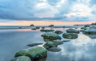 Southern coast of the Finnish Gulf. Rocks covered with green seaweed in the Baltic sea. Smooth transparent reflective water. Orange sunset markings under the low altitude clouds. Estonia, Baltic