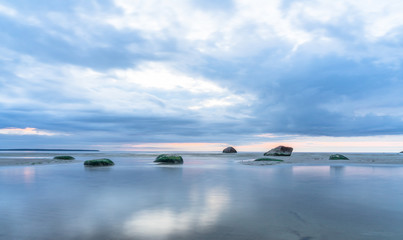 Southern coast of the Finnish Gulf. Rocks covered with green seaweed in the Baltic sea. Smooth transparent reflective water. Orange sunset markings under the low altitude clouds. Estonia, Baltic