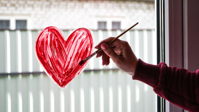 hands of a girl with a paintbrush painting red heart on a window, copy space for text, stay home