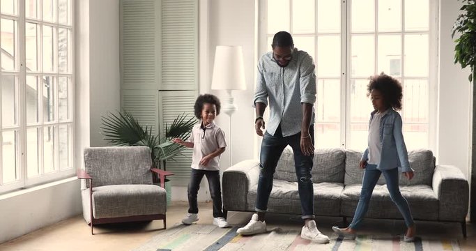Smiling african ethnicity father showing dancing moves to energetic little children in living room. Happy mixed race daddy giving dance class, teaching educating small daughter and son at home.