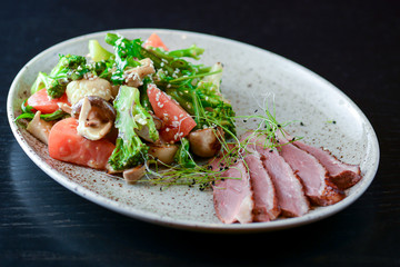 Green salad made with ham, tomatoes, mushrooms, broccoli, sesame seeds on a white plate, on dark wooden background.