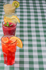 Three tropical mixed drinks, berries, orange and kiwi fruit cocktails served on green plaid tablecloth.
