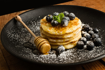 Tortitas con arándanos y hojas de menta con azúcar glass y miel sobre un plato redondo negro....