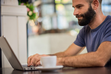 Young bearded Caucasian man sitting in restaurant and using laptop.