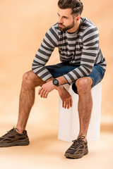 handsome stylish bearded man in striped sweatshirt sitting on white cube on beige