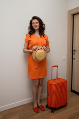 The girl holds a straw hat in her hands and looks away. She and a suitcase are standing near the front door.