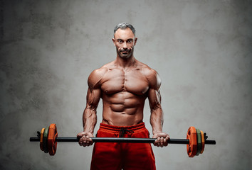 Shirtless adult male bodybuilder doing a biceps exercise with a barbell in a bright studio