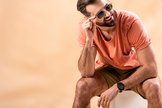 Happy Handsome Man In Shorts, Summer T-shirt And Sunglasses Sitting On White Cube On Beige