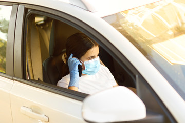 bambina con i capelli neri indossa una mascherina facciale e guanti in lattice per proteggersi e telefona dall'abitacolo di un automobile 