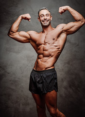 Shirtless adult male bodybuilder doing a biceps showing with a smile in a bright studio