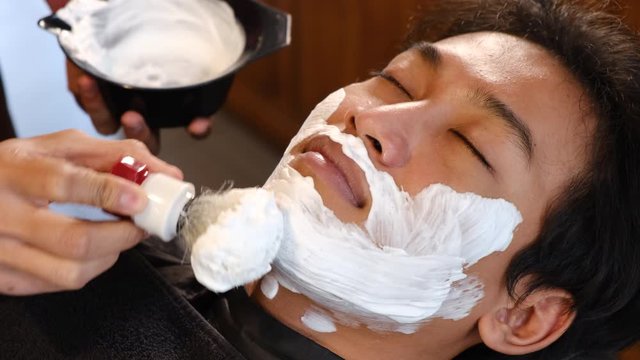 Professional hairdresser applying shaving foam to client's skin in barbershop. .Young asian man get his beard cut into shape in barbershop. Male beauty treatment concept