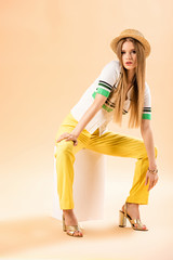 fashionable young woman in yellow trousers, polo and straw hat sitting on white cube on beige