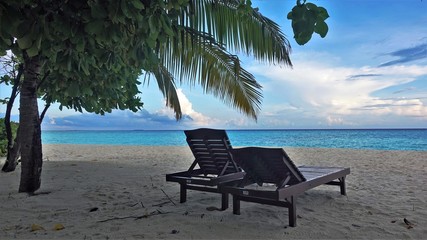 Maldives. On the clear white sand, under palm trees, there are sun loungers for relaxation. View of the azure ocean and blue sky with beautiful clouds.