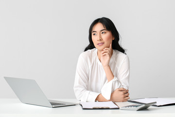 Beautiful Asian secretary at table against light background