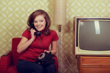 Young charming woman sitting in chair and talking on old phone in room with vintage wallpaper and retro TV set, retro stylization 60-70s 20th century. Girl in room with old fashioned interior