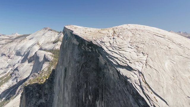 Impressive aerial shot of the top and the steep wall of the symbol of Yosemite National Park - the Half Dome rock, brightly lit with the sun. Sierra Nevada panorama at the background. 4K