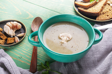 Close-up turquoise ceramic bowl with mushroom cream soup, wooden spoon, bread and garlic on grey napkin on green wooden background.