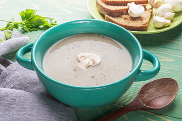 Turquoise ceramic bowl with mushroom cream soup, spoon, garlic, bread, grey napkin on green wooden background.