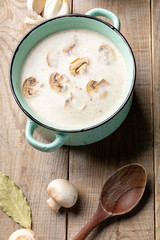 Turquoise ceramic pan with mushroom cream soup, fresh champignons, spoon, garlic and bay leafs on wooden background.