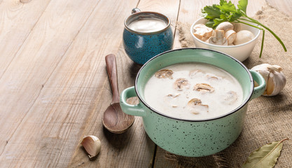 Turquoise ceramic pan with mushroom cream soup, sour cream, fresh champignons, spoon and spices on burlap on wooden background.