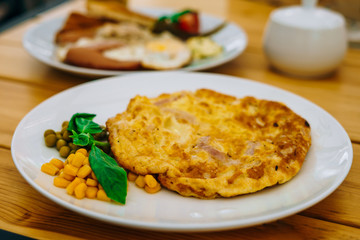 Omelet with ham, peas and corn in a cafe. Delicious omelet breakfast with cheese and peas. Omelet with vegetables on a white plate in a cozy cafe.