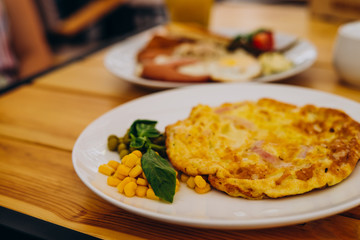 Omelet with ham, peas and corn in a cafe. Delicious omelet breakfast with cheese and peas. Omelet with vegetables on a white plate in a cozy cafe.