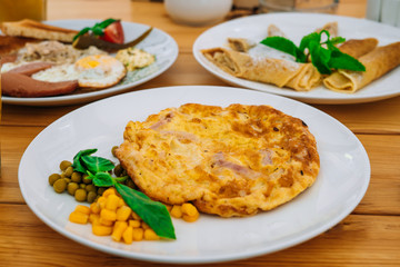 Omelet with ham, peas and corn in a cafe. Delicious omelet breakfast with cheese and peas. Omelet with vegetables on a white plate in a cozy cafe.