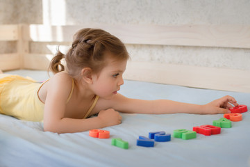 girl 3 years Caucasian in yellow dress lying on wooden bed on blue bedspread and learning in play...