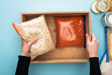 cropped view of woman packing groats in wooden box on blue background, food donation concept