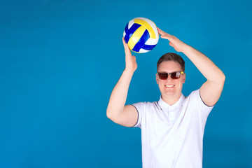 Naklejka premium A young volleyball fan, a man in a white shirt, stands with a ball in his hands on a blue background. The concept of recreation and travel.