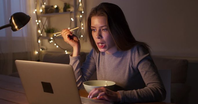Young woman working late on laptop and eating noodles