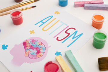 card with autism word and painting of head with brain on white with paint brushes, chalks and paints