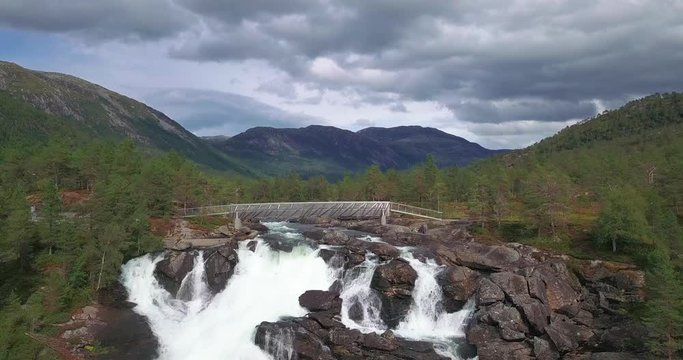 A narrow steel bridge spanning above the Likholefossen waterfall on the Gaula river in the Eldalsdalen valley. The river runs high, wild, and powerful. The wide valley covered with forest.