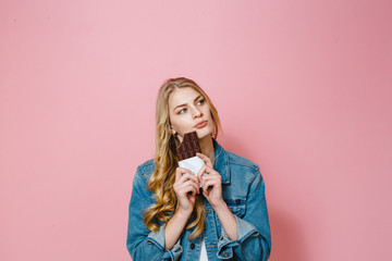 Portrait of a beautiful blonde girl eating chocolate is excited on a pink background