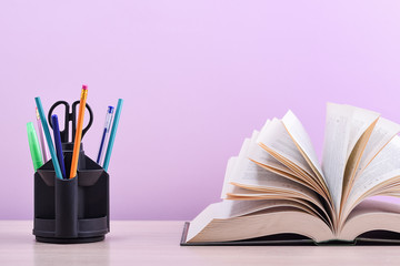 A large thick book with the pages spread out like a fan and a stand with pens, pencils and scissors on the table on a lilac background.