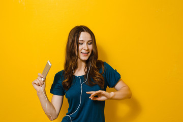 Portrait of a beautiful girl holding a cell phone,  and listen to music in the headphones over a yellow background