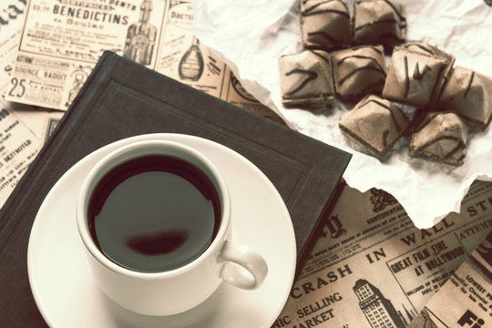LONDON, ENGLAND - APRIL 14, 2020. a cup of strong coffee on a white saucer and a black book on a table covered in old newspapers. cup with coffee and waffle cookies on white crumpled paper.