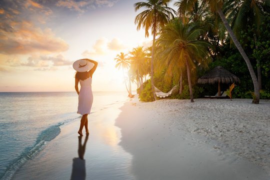 Eine attraktive Frau mit Hut steht am tropischen Strand unter Palmen auf den Malediven bei Sonnenuntergang