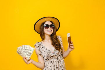 Image of a beautiful happy brunette girl with sunglasses isolated over yellow background holding money banknotes travel tickets and passport