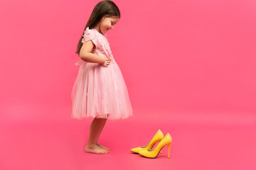 funny child girl fashionista in dress going to put on big mother's yellow shoes on colored background.