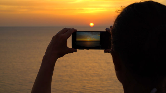Close up on woman's hands taking a photo with cell phone of beautiful sunset over the sea
