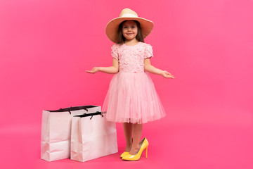 Happy lovely moments of shopping time with cute little girl in dress standing in mother`s big shoes with white packages in hands isolated on pink background