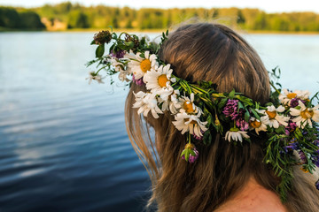 Blonde girl in a white dress with a flower wreath on her head is sitting on the pier. Scenic forest lake. Ligo. © Anastasia
