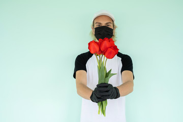 a guy in a white T-shirt with a black short sleeve and a white cap, in a black protective mask, is holding a bouquet of red tulip flowers in his hands Home delivery during quarantine